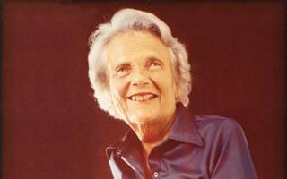 Katherine Neuberger smiling in shiny dark colored blouse with black background 