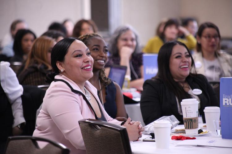 Women of different ages and races smile as they sit at tables in a conference room