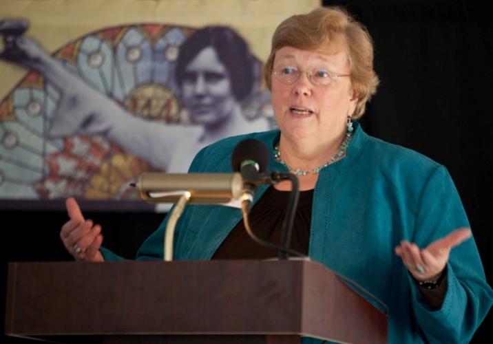 Susan Carroll in teal jacket and black top, speaking at podium