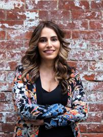 Head shot Senator Teresa Ruiz wearing a multi-color blazer and black shirt, standing with arms crosses against a brick background